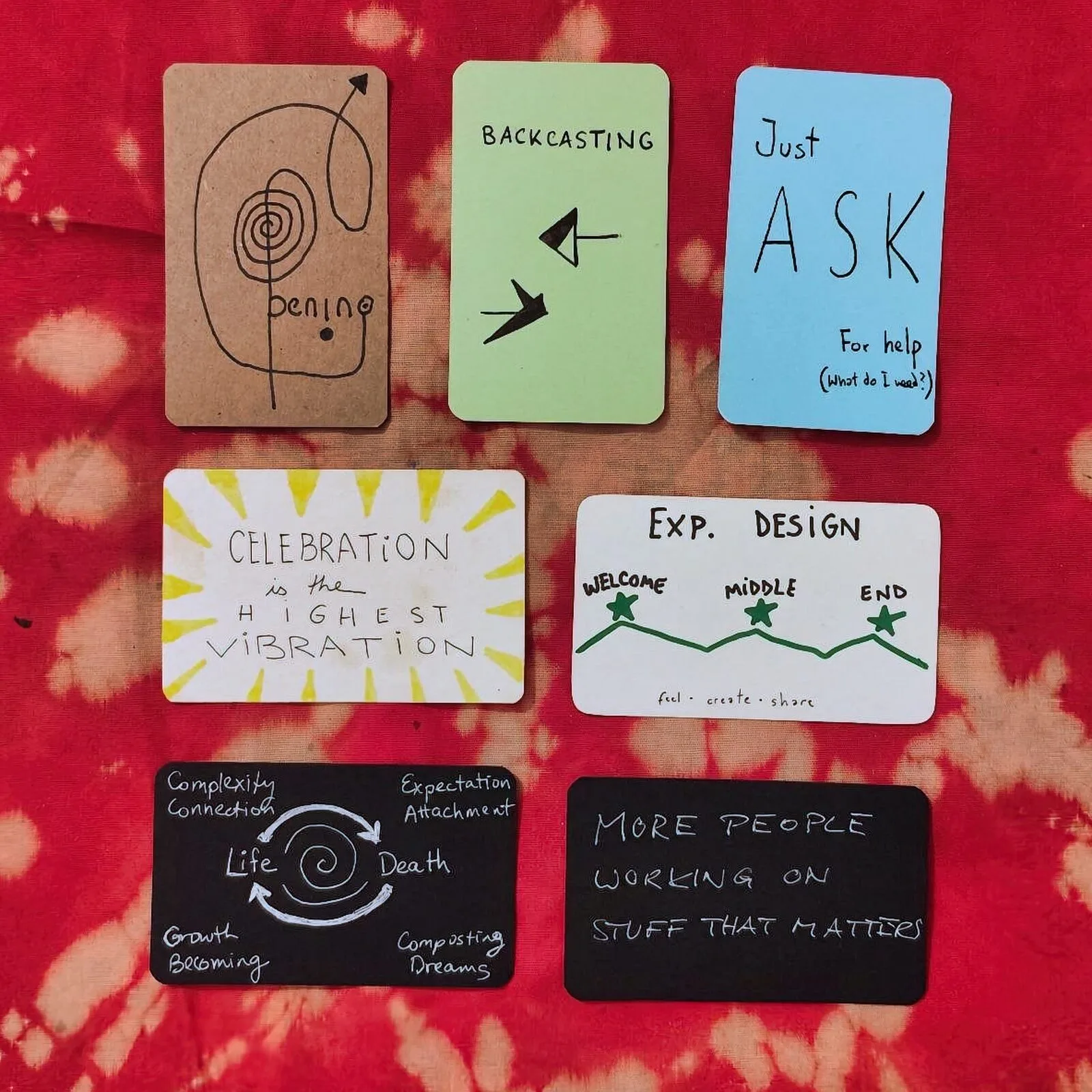 Seven hand-drawn prototype cards laid out on red tie-dyed cloth: Beginning, Backcasting, Just Ask, Celebration is the highest vibration, Exp. Design, a life and death cycle, and More people working on stuff that matters
